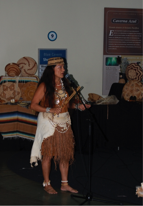 Woman in basket hat, abalone-shell necklaces and clothing of grass and rawhide beats out rhythm with clapper stick; display of baskets in background