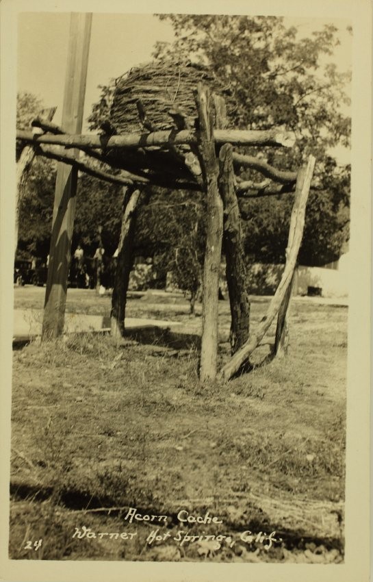 Sepia-toned photograph of log framework supporting beehive-shaped storage container made of sticks