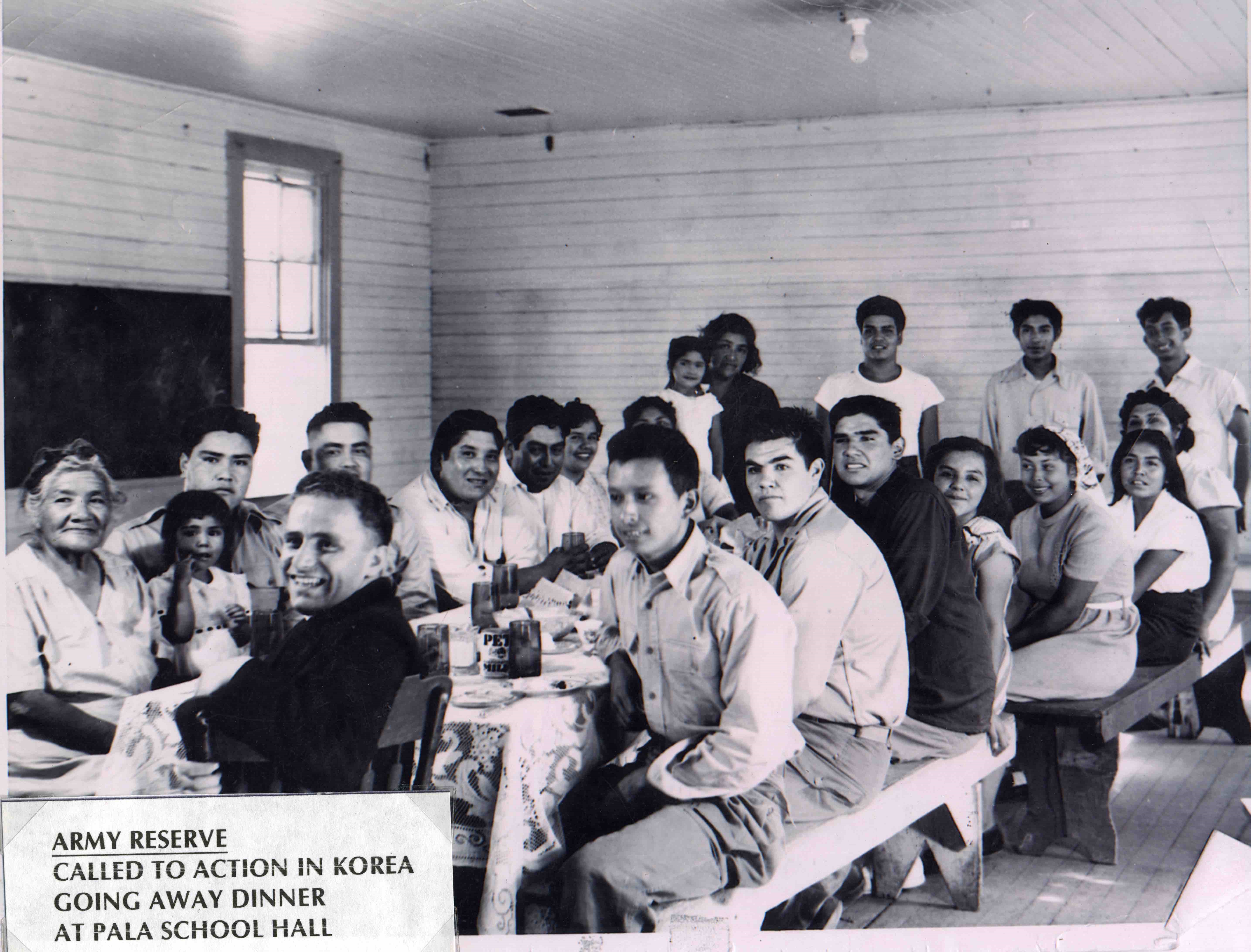 Black-and-white photograph of young men in U.S. Army uniforms with elders, children and other relatives sitting down to eat at long tables in a dining hall; printed text that reads: “ARMY RESERVE: CALLED TO ACTION IN KOREA: GOING AWAY DINNER AT PALA SCHOOL HALL”