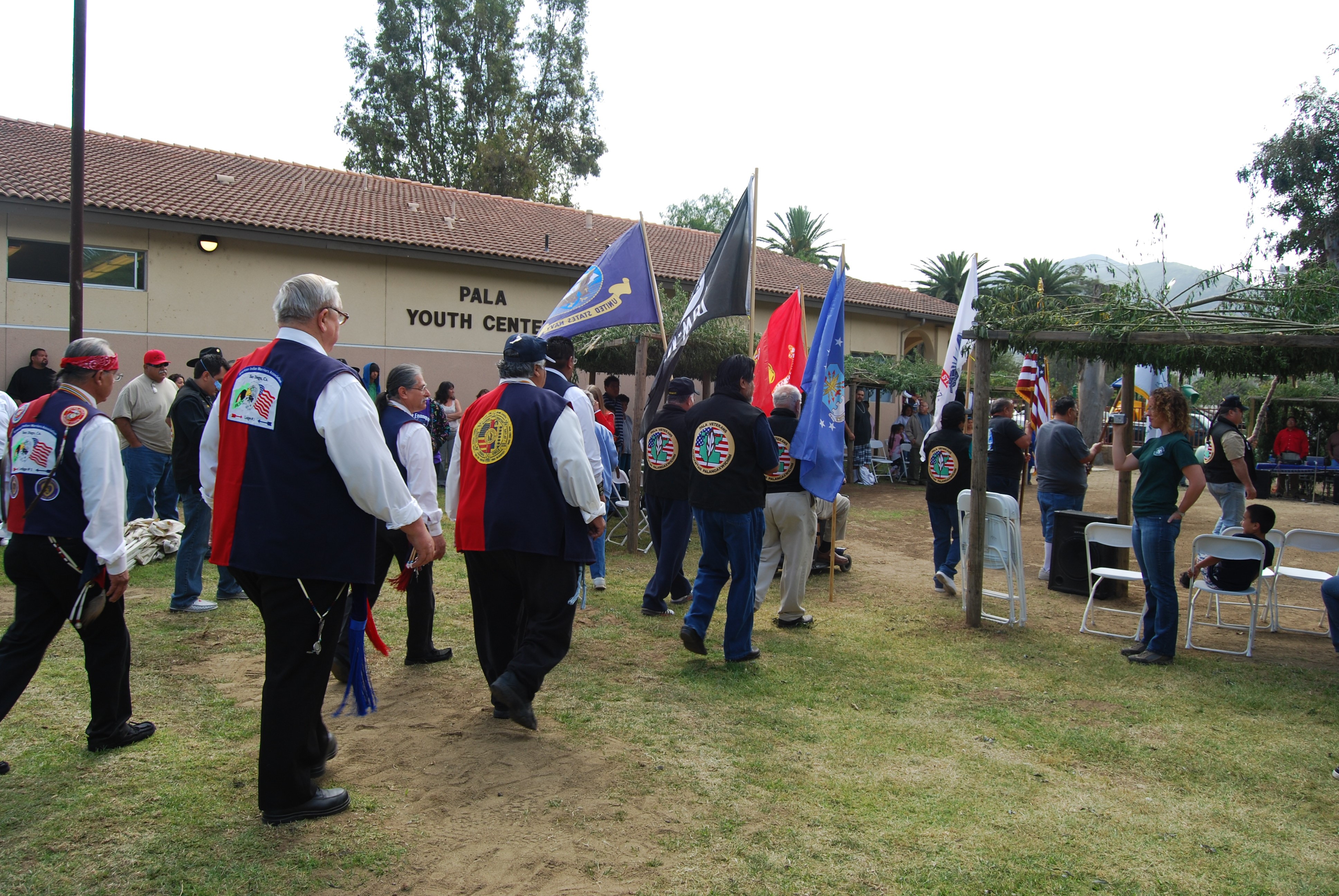 A procession of men in jackets and vests representing their tribal and veteran status escorting tribal flags past traditional ramadas (arbors) toward the Pala Youth Center