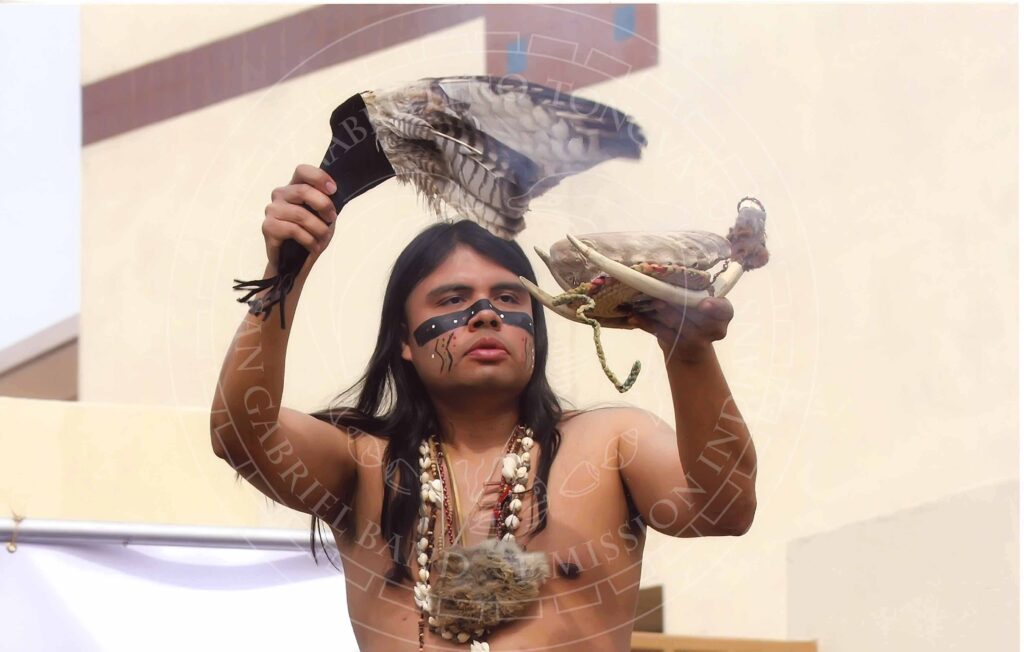 Young man with long hair in traditional regalia including bead and shell necklaces and black face paint with red and white dots fans an incense holder made of abalone shell and other natural materials with a fan made of feathers