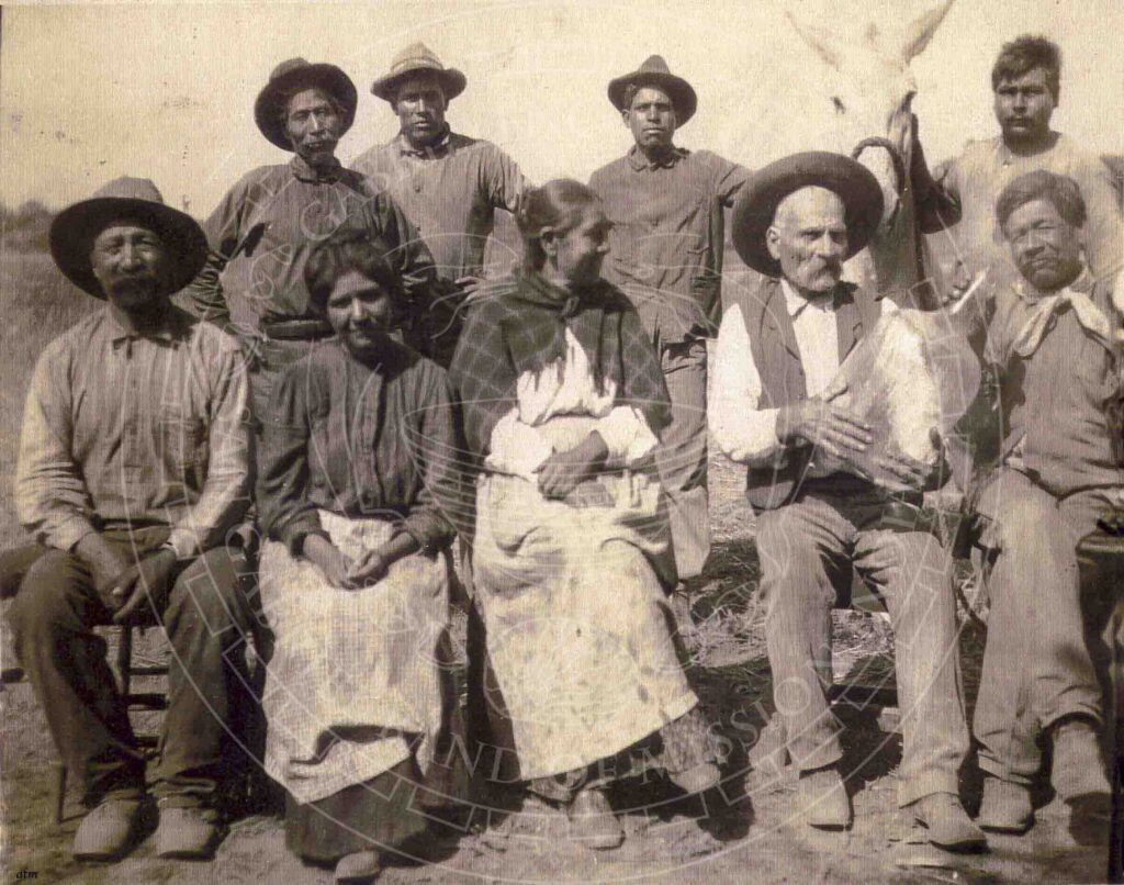 Black and white historic image of men and women in hats, bandanas, vests, boots and other items of Western wear posing seated in chairs with another row standing behind them. At right, two seated men hold a large glass bottle while two standing men behind them hold onto a white horse.