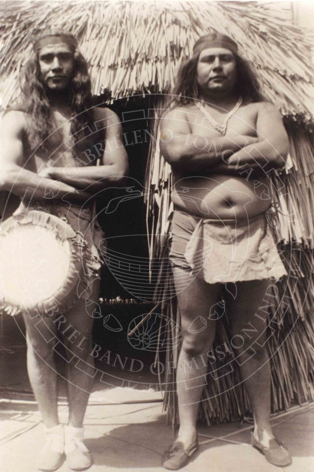 Sepia toned historical photograph of two long-haired men in traditional attire including headbands, aprons and necklaces stand with their arms crossed in front of a thatched house