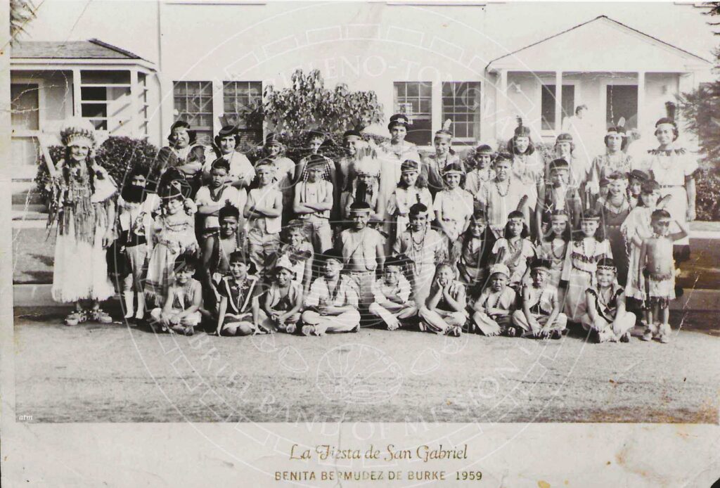 Sepia toned historic image of a large group of children and adults wearing Native costumes seated and standing in front of a white bulding. Text reads "La Fiesta de San Gabriel, Benita Bermudez De Burke, 1959"
