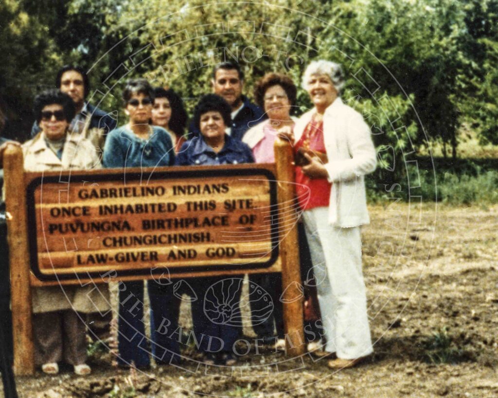 Color photograph of men and women in modern attire standing around a sign that reads: "Gabrielino Indians once inhabited this site, Puvungna, birthplace of Chungichnish, Law-Giver and God."