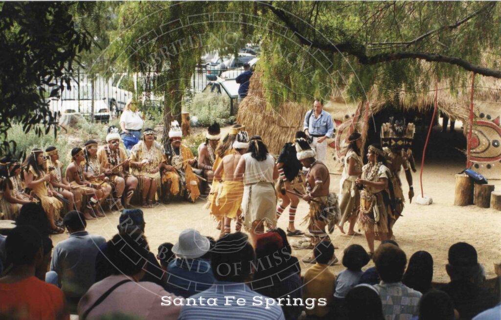 A circle of people in both traditional and modern attire including headdresses, necklaces, aprons and grass skirts watch a group of dancers in regalia performing in front of a large thatched house with painted decorations around the doorway.
