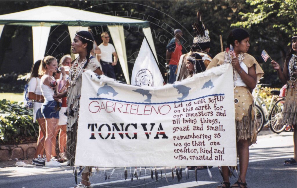 Two young women in traditional attire including hide dress and bead necklaces march in parade with a banner that reads: "Gabrielino Tongva: Let us walks softly on this earth for our ancestors and all living things, great and small, remembering as we go that one creator, kind and wise, created all.