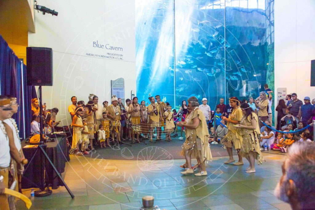 Procession of three women in traditional attire including hide dresses, necklaces and headbands approach a table flanked by others in traditional dress. They are in an aquarium with an enormous fish tank, with the title "Blue Cavern: Presented by American Honda Motor Co., Inc."