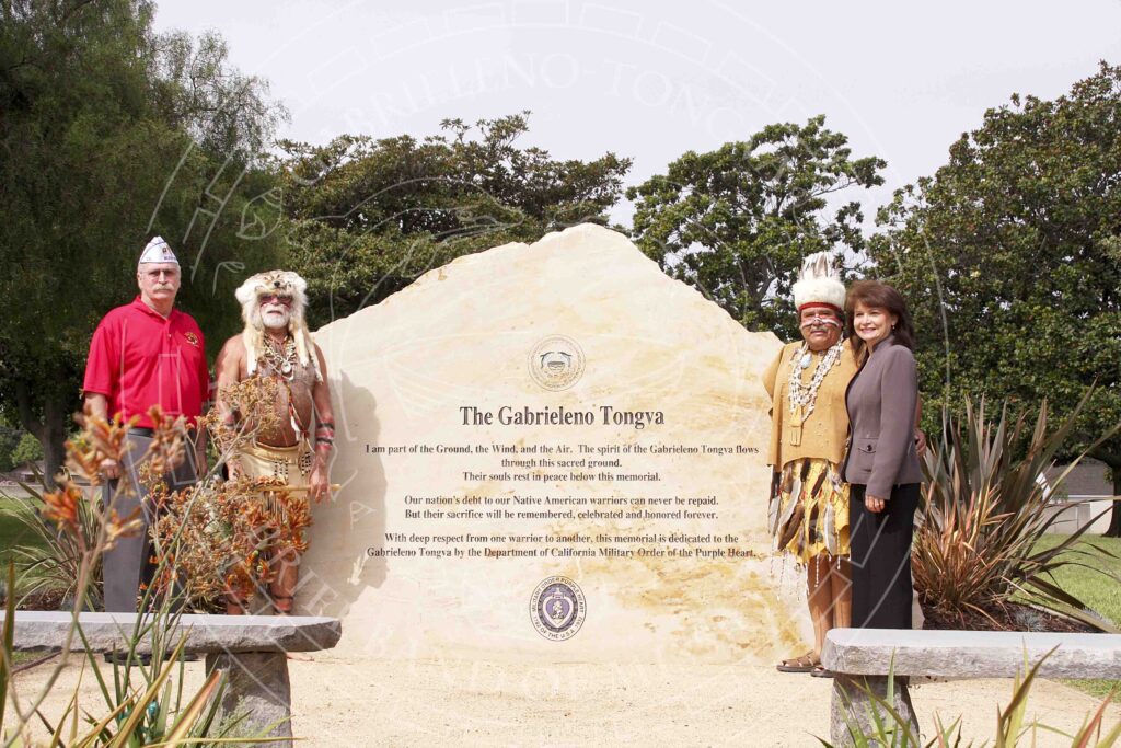 Adults in a mix of traditional and modern attire including veteran's forage cap and fur and feather headdresses standing next to a large carved stone with an inscription reading: "The Gabrielleno Tongva: I am part of the Ground, the Wind, and the Air. The spirit of the Gabrieleno Tongva flows through this sacred ground. Their souls rest in peace below this memorial. Our nation's debt to our Native American warriors can never be repaid. But their sacrifice will be remembered, celebrated and honored forever. With deep respect from one warrior to another, this memorial is dedicated to the Gabrieleno Tongva by the Department of California Military Order of the Purple Heart." Above: Tribal seal; below: seal of the Order of the Purple Heart.