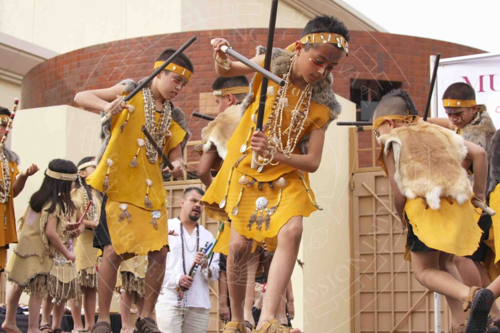 Children dancing in traditional attire including headbands, necklaces, furs, and hide clothing with shell and bead decorations. Boys in foreground are using clapper sticks to beat out a rhythm while they dance.