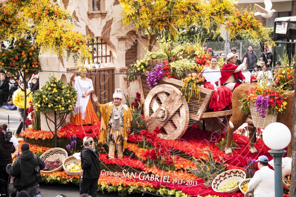 Parade float designed to look like a mission building with fruit trees and a donkey cart loaded with baskets of flowers. Men and women on the float wear a mix of traditional Tongva and Hispanic or Mexican attire.