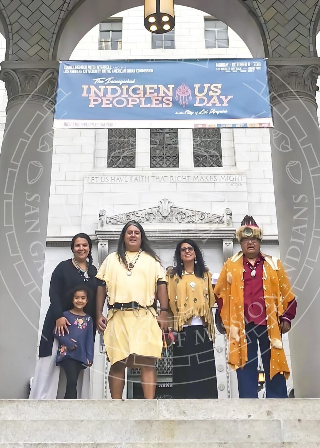 Adults and children in a mix of traditional and modern attire standing in front of an entrance to a neoclassical public building with a banner celebrating the inaugural Indigenous Peoples Day for the City of Los Angeles