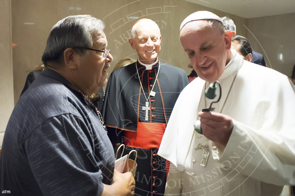 Pope and Vatican official in traditional Catholic robes laughing as they converse with a man in modern attire with a Tribal necklace. Pope is holding a small bronze bell.