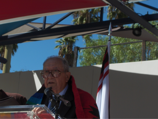 Man in brightly-colored shawl speaking at podium in front of Gabrielino/Tongva flag