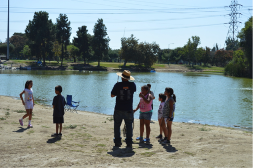 Alt-text: Man demonstrating fishing pole to children on shore of lake in park