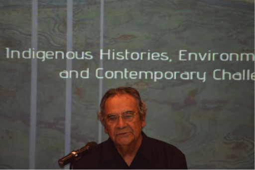 Man speaking into microphone in front of slide reading (in part) “Indigenous Histories”