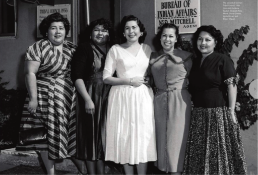 Black-and-white photo of five women in elegant 1950s dresses standing in front of a building with signs reading “Bureau of Indian Affairs, Ned Mitchell, Agent” and (partly obscured) Tribal Council 1965