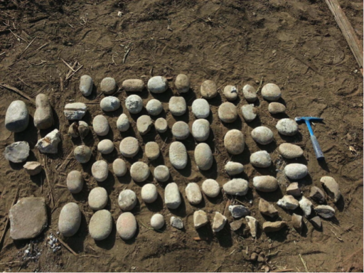 A collection of stone tools, mostly round-edged, flat stones, laid out in dirt beside an archaeologist’s hammer