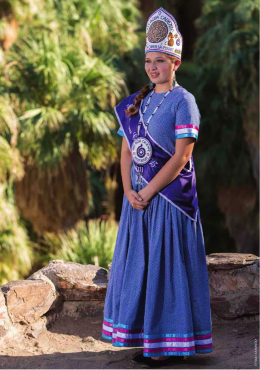 Photo of smiling young woman in purple costume with embroidered conical hat, sash, beadwork pendant and shimmering stripes on the lower sleeves and skirt, posing in desert landscape with rocks and palm trees
