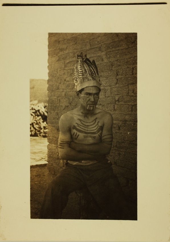 Sepia-toned photograph of a man with crossed arms in feathered headdress and body decorations