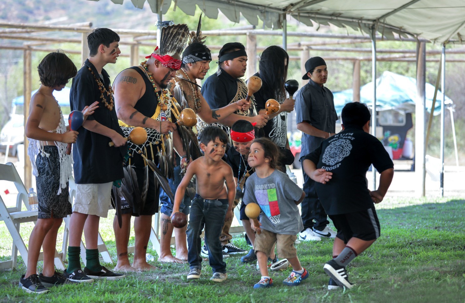 Young men and boys dancing and shaking gourd rattles in a mix of traditional regalia and modern dress