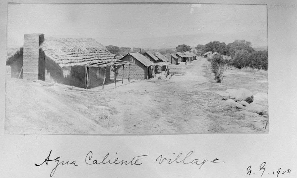 Old photograph of a neat row of houses with adobe walls and thatched roofs along a dirt road