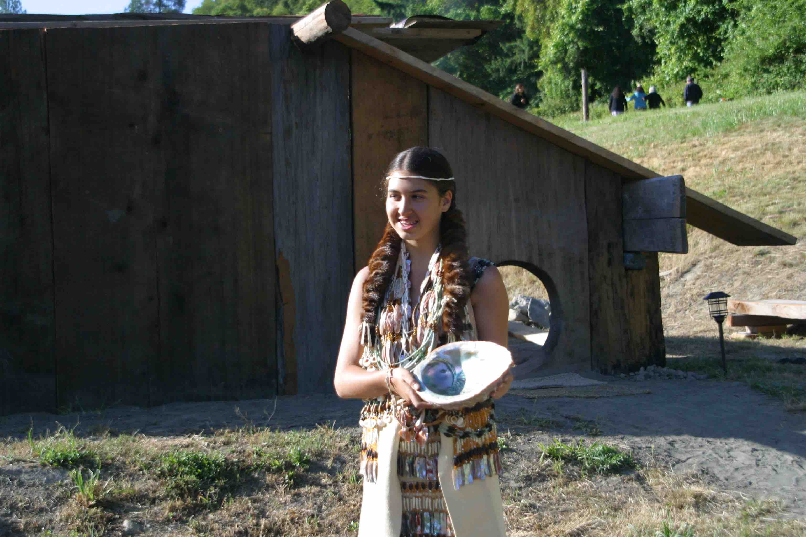 A young woman in traditional regalia including beaded vest and necklaces holding abalone shell in front of wooden house with round opening