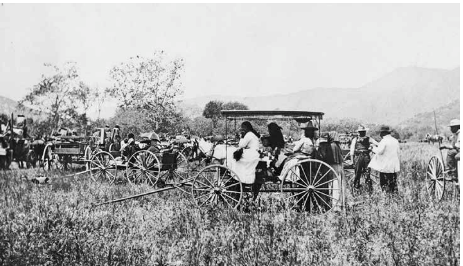 Old photograph of families waiting in and around unhitched wagons in an overgrown field with mountains in background