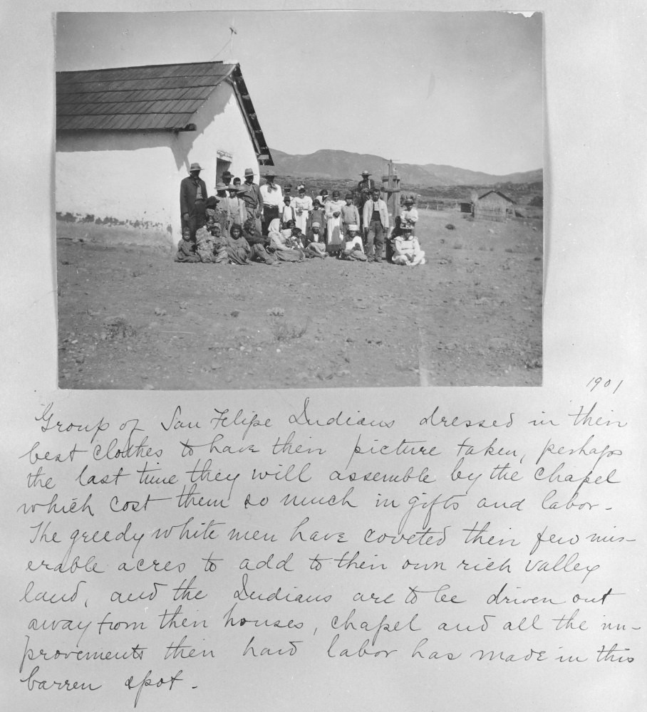 Old photograph of men, women and children sitting and standing in front of an adobe building with handwritten text that reads: “1901: Group of San Felipe Indians dressed in their best clothes to have their picture taken, perhaps the last time they will assemble by the chapel which cost them so much in gifts and labor. The greedy white men have coveted their few miserable acres to add to heir own rich valley land, and the Indians are to be driven out away from their houses, chapel and all the improvements their hard labor has made in this barren spot.”