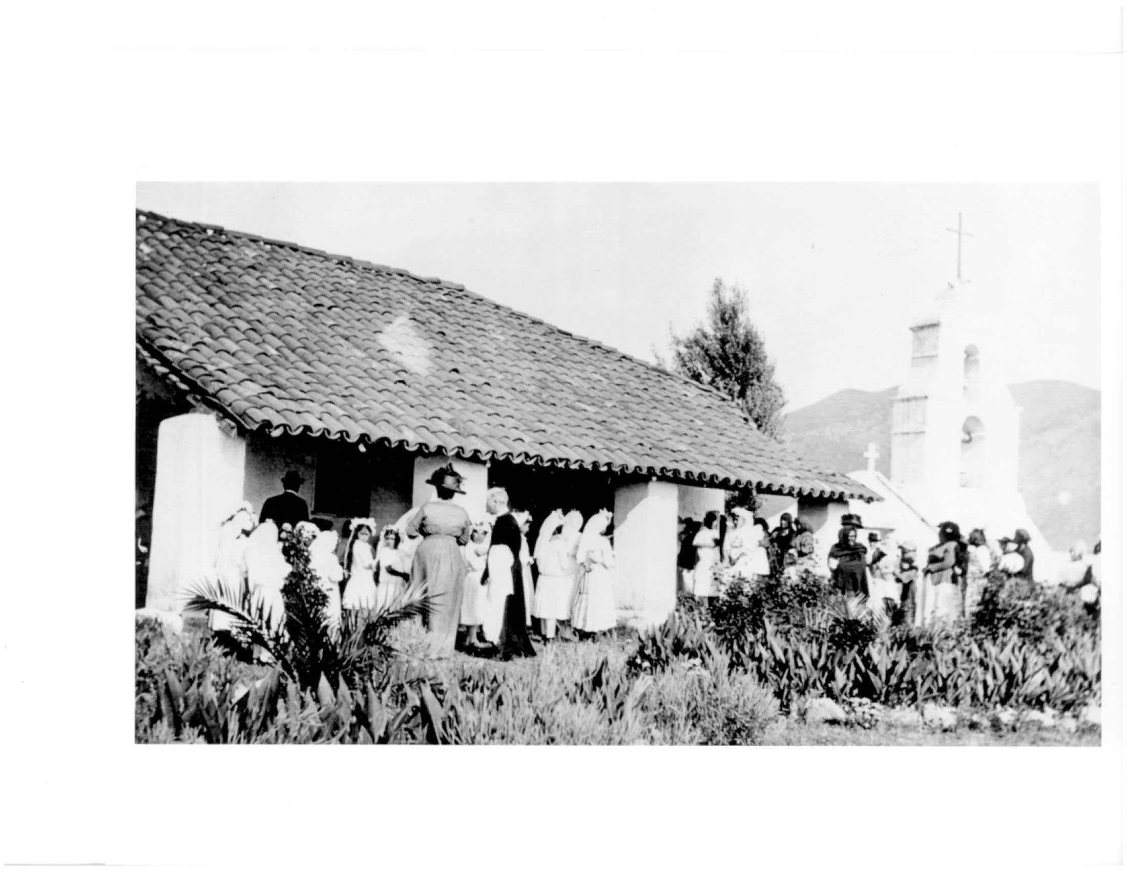 Old photograph of people in white dresses and other finery in front of tile-roofed adobe surrounded by agave plants with belfry in background