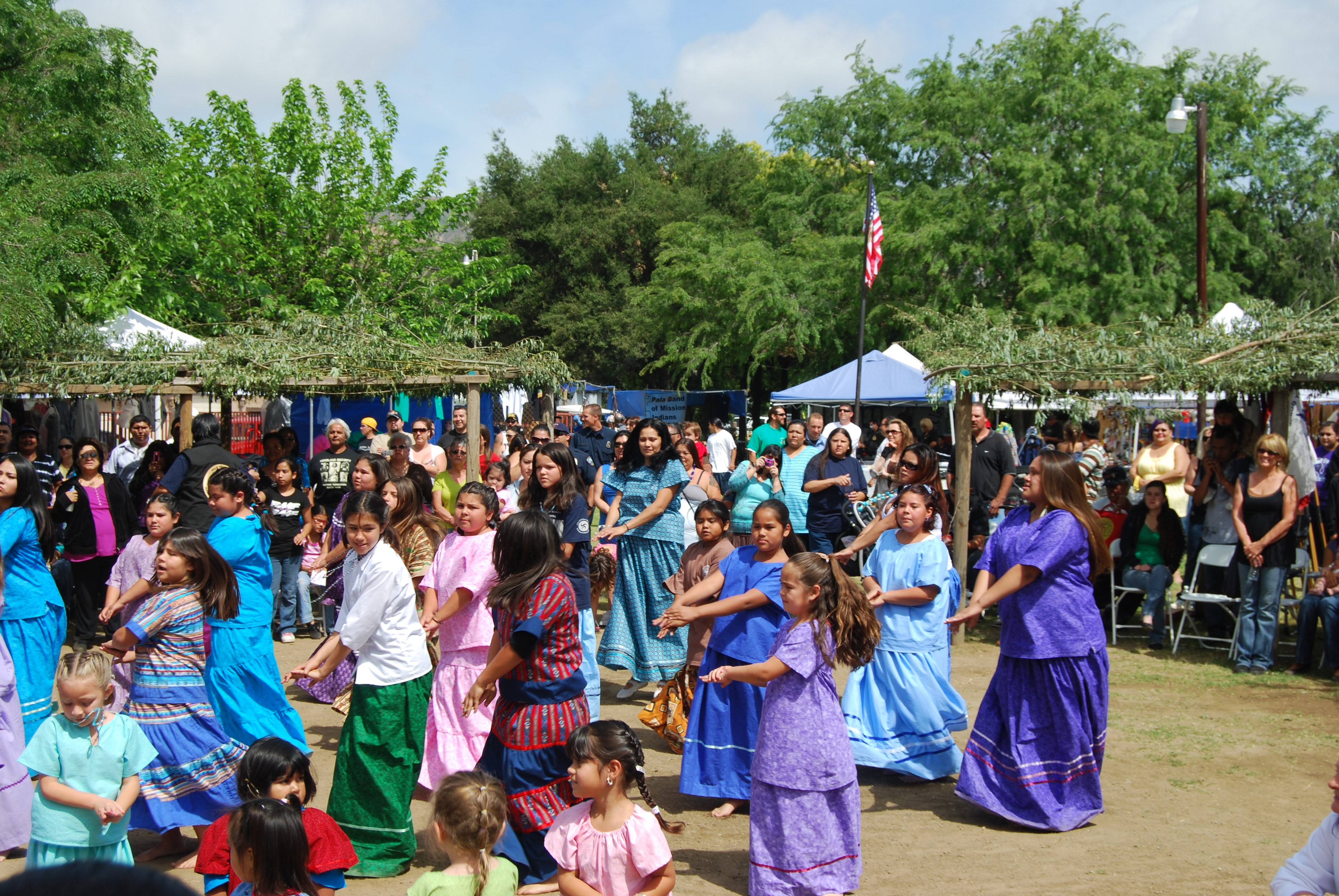 Women and girls in colorful dresses dancing in a circle surrounded by traditional ramadas (awnings made of foliage) and modern festival booths