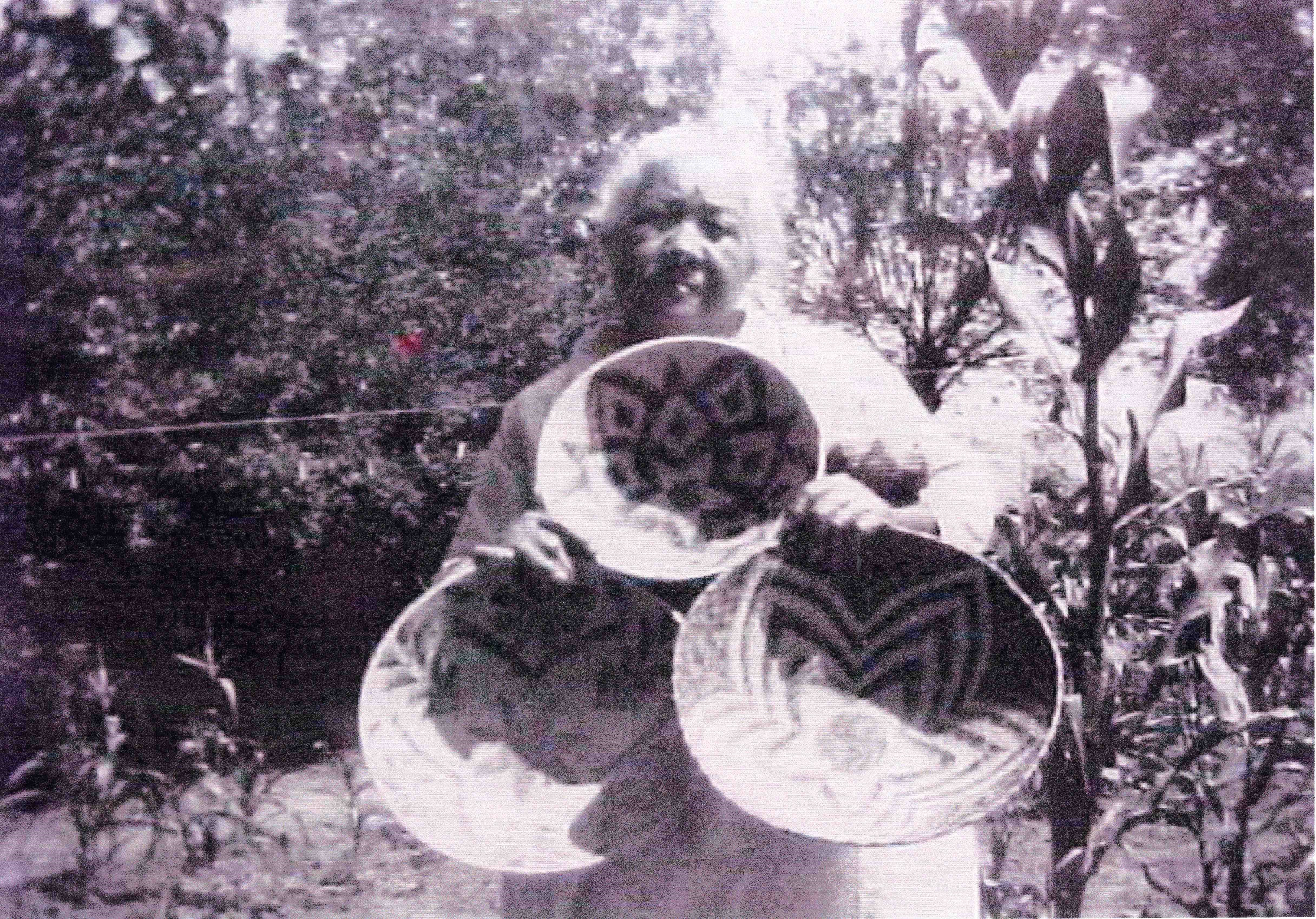 Black-and-white photograph of woman holding up three patterned baskets in front of a small patch of corn