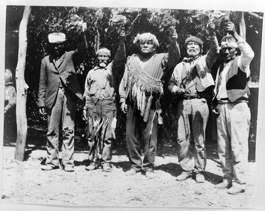 Old photograph of men wearing a mix of Western clothing and traditional regalia including headdresses and feathered sashes hold up rattles decorated with feathers