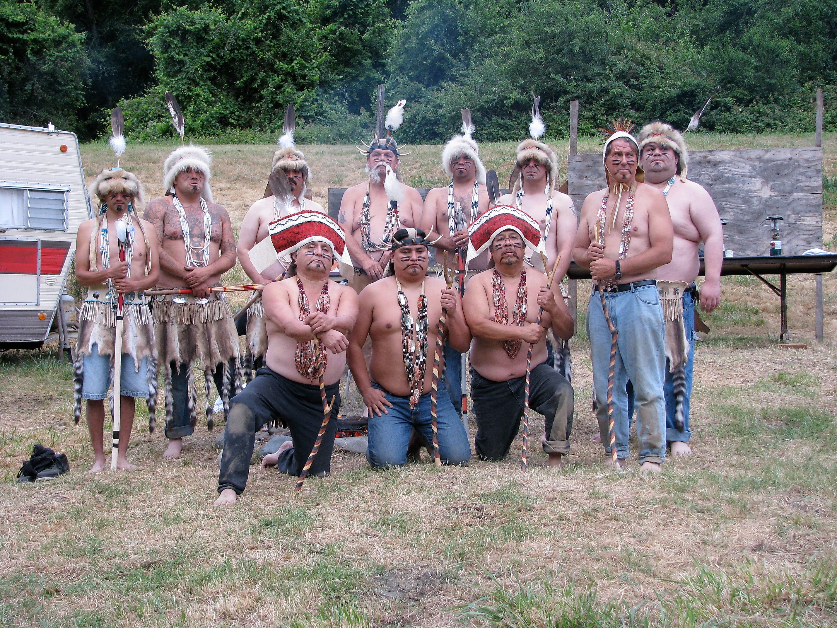 Eleven men in tribal regalia including headbands, bead necklaces, hide aprons and dance sticks standing and kneeling in a meadow