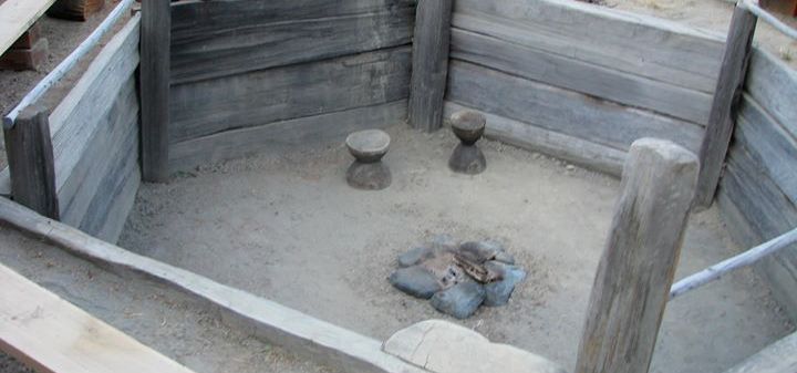 A submerged octagonal space with wooden plank walls and a dirt floor with a stone firepit and stools